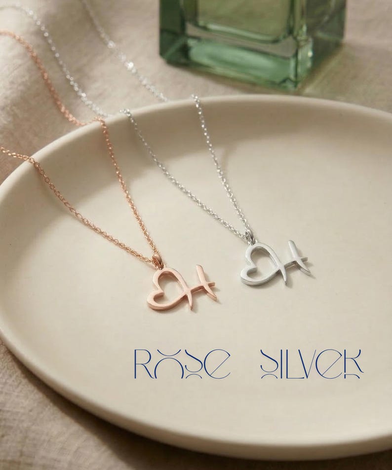 May include: Two delicate necklaces with heart-shaped pendants and the letter "H". One is rose gold, the other silver. The necklaces are displayed on a white plate. The text "ROSE SILVER" is below the necklaces.