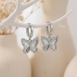 May include: A pair of silver hoop earrings with butterfly pendants. Each butterfly pendant is detailed with clear crystals around the edges and a textured design on the wings. The earrings are displayed on a white, branch-like structure.