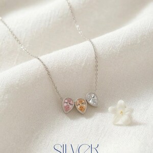 May include: A silver necklace with three teardrop-shaped pendants in pink, orange, and clear gemstones. The word "SILVER" is displayed in a stylized font at the bottom of the image. A small white flower is next to the necklace.