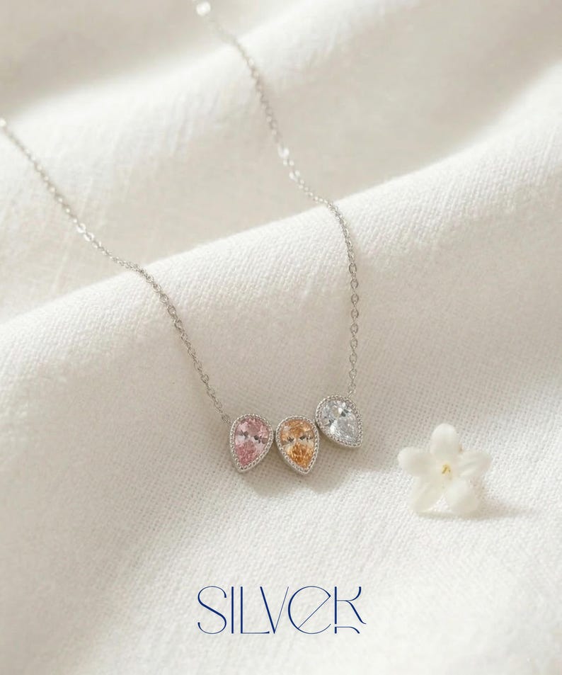 May include: A silver necklace with three teardrop-shaped pendants in pink, orange, and clear gemstones. The word "SILVER" is displayed in a stylized font at the bottom of the image. A small white flower is next to the necklace.