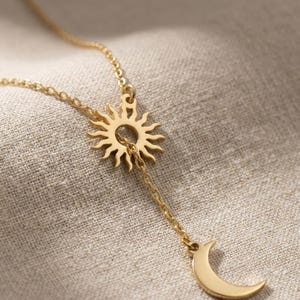 May include: Gold-colored necklace featuring a sun pendant with a dangling crescent moon charm. The sun pendant has a detailed, radiant design. The necklace is displayed on a neutral-toned fabric background, highlighting the jewelry's warm hue.