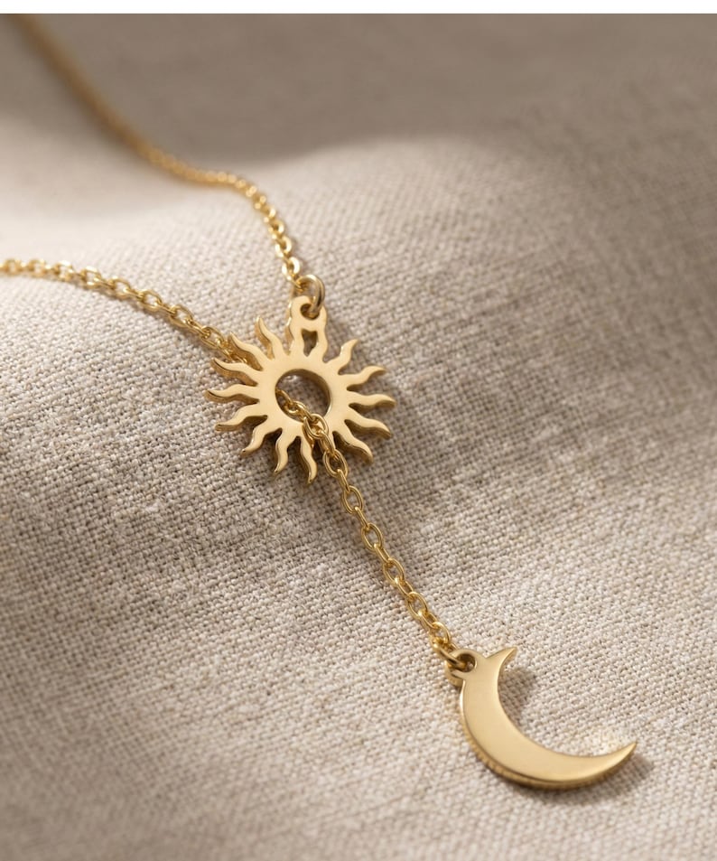 May include: Gold-colored necklace featuring a sun pendant with a dangling crescent moon charm. The sun pendant has a detailed, radiant design. The necklace is displayed on a neutral-toned fabric background, highlighting the jewelry's warm hue.