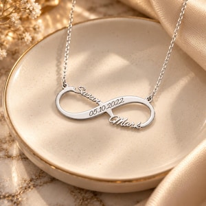 Op de afbeelding: Een zilverkleurige ketting met een oneindigheidssymbool, gegraveerd met de namen Susan en Mark en de datum 05.10.2022. De hanger is bevestigd aan een fijne zilverkleurige ketting en heeft een gepolijste metalen afwerking met cursief schrift.