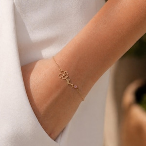 May include: A delicate gold bracelet with a floral design and a small pink gemstone. The bracelet is worn on a wrist, highlighting its intricate details and elegant simplicity. The bracelet's chain is thin and the flower is a cutout design.