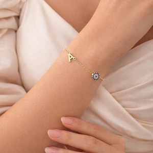 May include: A gold-colored bracelet featuring the letter "A" and an eye-shaped charm with blue and white accents. The bracelet is delicate and worn on a wrist.