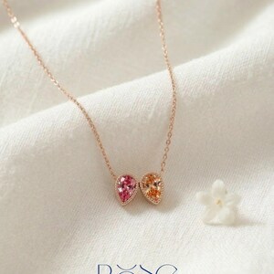 May include: A rose gold necklace featuring two teardrop-shaped pendants, one pink and one orange, set with small gemstones. The necklace is displayed on a white fabric with a small white flower and the word "ROSE" in blue.