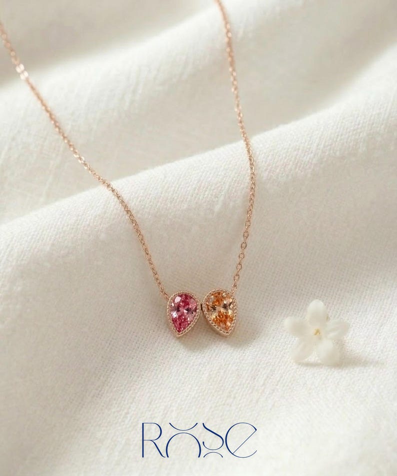 May include: A rose gold necklace featuring two teardrop-shaped pendants, one pink and one orange, set with small gemstones. The necklace is displayed on a white fabric with a small white flower and the word "ROSE" in blue.
