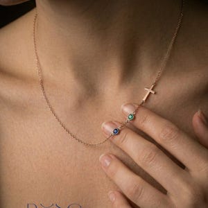 Puede incluir: Un delicado collar de oro rosa con un colgante de cruz y dos peque&ntilde;as piedras preciosas redondas en azul y verde. El collar se muestra en el cuello de una persona, mostrando su elegante dise&ntilde;o. La palabra "ROSE" est&aacute; abajo.