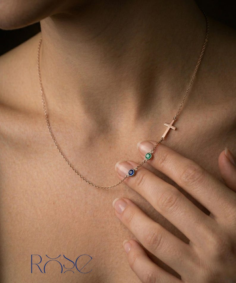 Puede incluir: Un delicado collar de oro rosa con un colgante de cruz y dos peque&ntilde;as piedras preciosas redondas en azul y verde. El collar se muestra en el cuello de una persona, mostrando su elegante dise&ntilde;o. La palabra "ROSE" est&aacute; abajo.