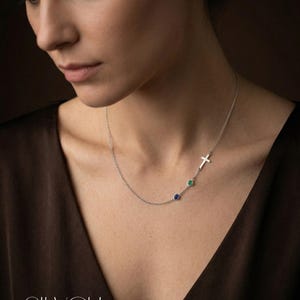 Puede incluir: Un collar de plata con un colgante de cruz y dos gemas de colores. El collar tiene una cadena fina y lo lleva una persona. Las gemas son azules y verdes. La palabra "SILVER" est&aacute; en la parte inferior de la imagen.