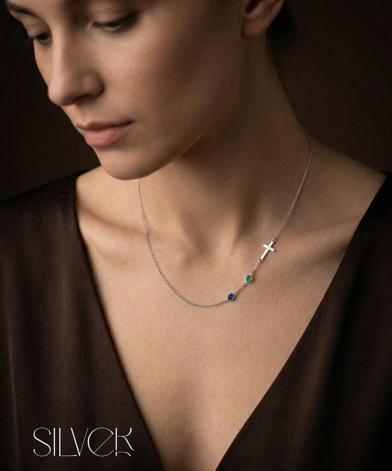 Puede incluir: Un collar de plata con un colgante de cruz y dos gemas de colores. El collar tiene una cadena fina y lo lleva una persona. Las gemas son azules y verdes. La palabra "SILVER" est&aacute; en la parte inferior de la imagen.