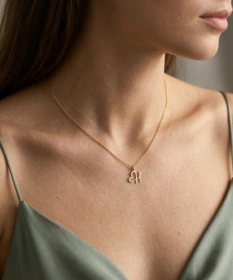 May include: A delicate gold necklace with a pendant shaped like the letter "H" intertwined with a heart. The necklace is worn against a person's skin, and the chain is thin and elegant. The background is a soft, neutral tone.