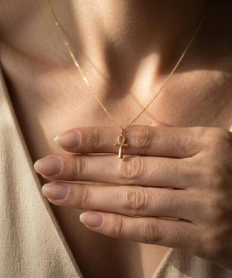 May include: A delicate gold-colored necklace featuring an Ankh pendant. The pendant is suspended from a fine chain. The necklace is displayed against a neutral background, highlighting the jewelry's design and color.