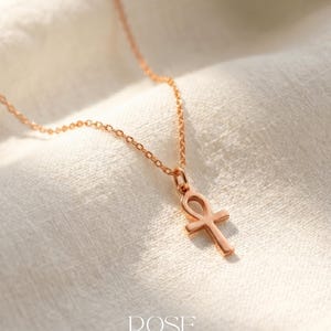 May include: Rose gold ankh pendant necklace. The ankh, a symbol of life, hangs from a delicate chain. The necklace is displayed on a white, textured fabric. The word "ROSE" is printed at the bottom.