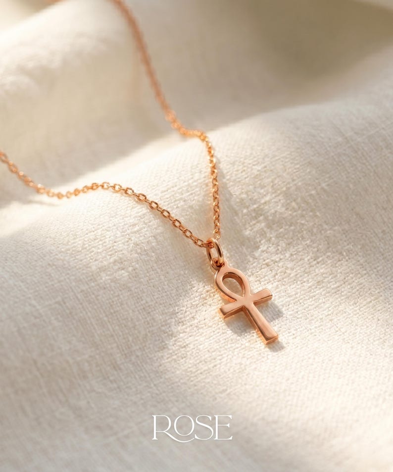 May include: Rose gold ankh pendant necklace. The ankh, a symbol of life, hangs from a delicate chain. The necklace is displayed on a white, textured fabric. The word "ROSE" is printed at the bottom.