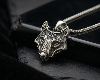Pode incluir: Um pingente de metal prateado em forma de cabeça de lobo com padrões gravados intrincados nas orelhas e na testa. O pingente está preso a uma corrente de metal prateado fina e trançada que se estende para cima a partir da cabeça.
