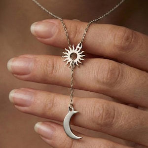 May include: A silver-toned necklace featuring a sun pendant at the top and a crescent moon charm suspended below. The sun pendant has radiating rays, and the moon is a smooth, curved shape. The necklace is displayed against a blurred background.