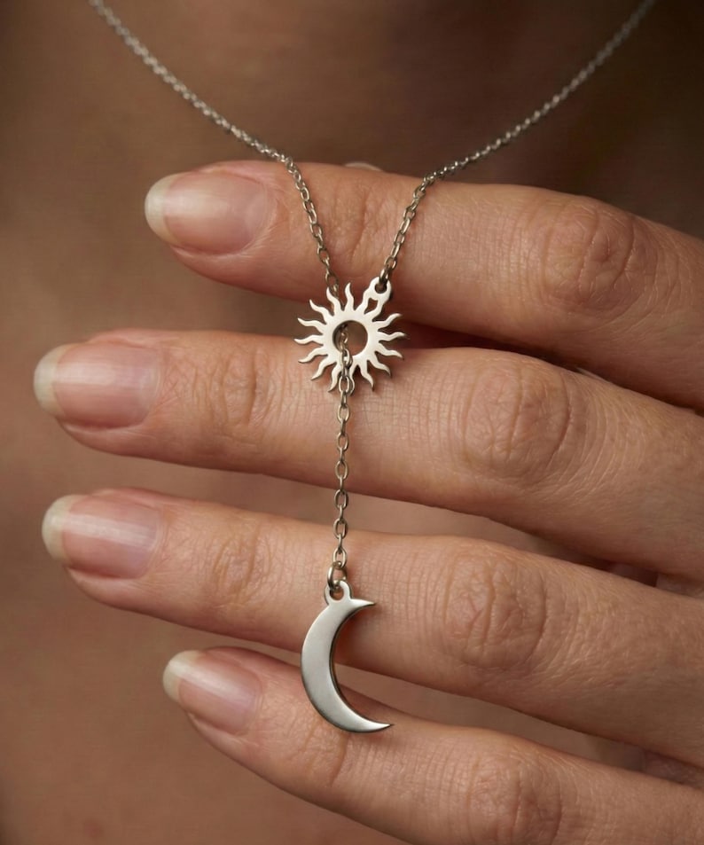 May include: A silver-toned necklace featuring a sun pendant at the top and a crescent moon charm suspended below. The sun pendant has radiating rays, and the moon is a smooth, curved shape. The necklace is displayed against a blurred background.