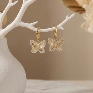 May include: A pair of gold-colored butterfly hoop earrings. Each earring features a butterfly pendant with a detailed wing design, outlined with small, sparkling crystals. The earrings are suspended from gold hoops.