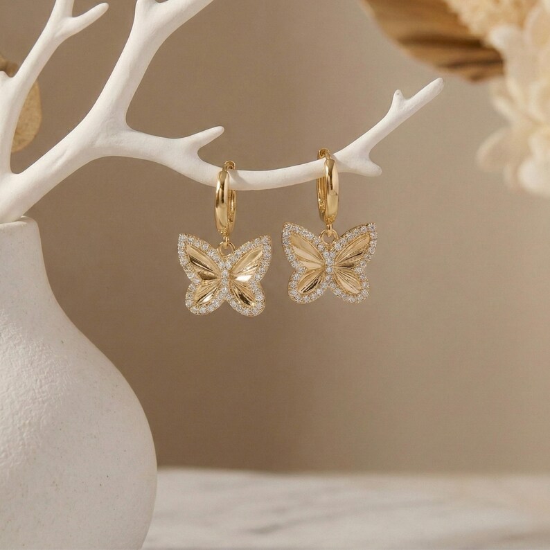 May include: A pair of gold-colored butterfly hoop earrings. Each earring features a butterfly pendant with a detailed wing design, outlined with small, sparkling crystals. The earrings are suspended from gold hoops.