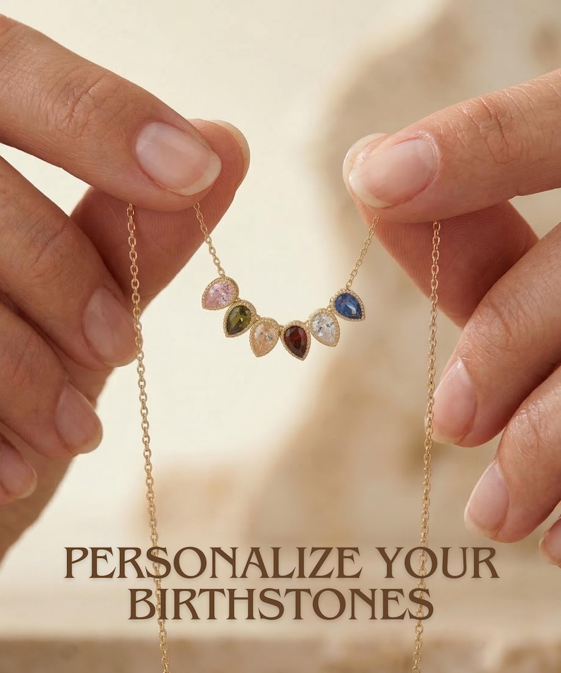 Könnte beinhalten: Eine goldene Halskette mit einer Kette und einer Reihe von tropfenförmigen Edelsteinen in verschiedenen Farben. Die Halskette wird von Händen gehalten, und der Text "PERSONALIZE YOUR BIRTHSTONES" wird darunter angezeigt. Die Edelsteine sind rosa, grün, pfirsichfarben, rot, weiß, blau.