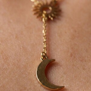May include: Gold-colored necklace featuring a sunburst pendant and a crescent moon charm. The necklace has a delicate chain and is designed to be worn around the neck. The sunburst pendant is at the top, with the moon charm hanging below.
