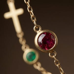 Puede incluir: Primer plano de un collar de oro con un colgante de piedras preciosas de rub&iacute; y esmeralda. El collar tiene una cadena delicada y un colgante de cruz en el fondo. El rub&iacute; es de color rojo intenso y la esmeralda es verde.