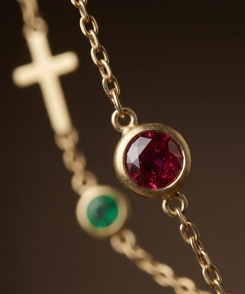 Puede incluir: Primer plano de un collar de oro con un colgante de piedras preciosas de rub&iacute; y esmeralda. El collar tiene una cadena delicada y un colgante de cruz en el fondo. El rub&iacute; es de color rojo intenso y la esmeralda es verde.
