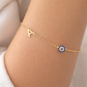 May include: Gold-colored bracelet featuring the letter "A" and an evil eye charm. The evil eye charm has a blue and white design. The bracelet is worn on a wrist.