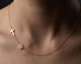 Pode incluir: Um colar de tom dourado com uma corrente fina e dois pingentes assimétricos: uma cruz pequena e simples e um motivo floral vazado, posicionados lado a lado na região da clavícula.