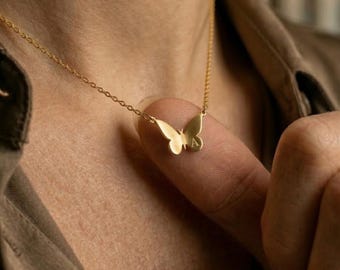 Puede incluir: Un delicado collar con colgante de mariposa dorada. El dije de mariposa está detallado con una textura sutil y presenta la letra "A". El collar se muestra en el cuello de una persona, mostrando el diseño y la cadena del colgante.