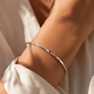Può includere: Un bracciale rigido in metallo color argento sottile con una superficie martellata e testurizzata. Il bracciale è di forma circolare e presenta una singola piccola pietra rotonda di colore scuro incastonata al centro della fascia.