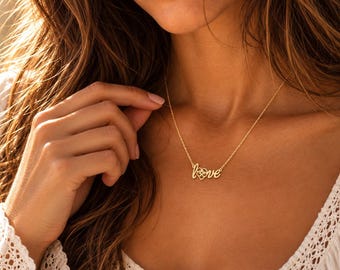 Pode incluir: Um colar de tom dourado com uma corrente fina e um pendente que forma a palavra love em letra cursiva. A letra o é substituída por um pequeno coração com um design floral recortado no centro, posicionado sobre a zona do pescoço.