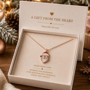 Könnte beinhalten: Eine roségoldfarbene Halskette mit einem Weihnachtsmann-Anhänger aus weißem Emaille mit Brille, präsentiert in einer cremefarbenen Geschenkbox. Der Deckel trägt die Aufschrift A GIFT FROM THE HEART und innen steht MADE WITH LOVE und weiterer Text.