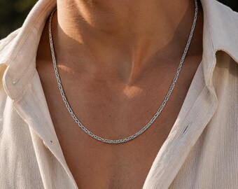 Puede incluir: Un collar de cadena plateada con un diseño tejido. El collar se lleva sobre una camisa abotonada de color claro. La cadena es un accesorio sencillo y elegante.