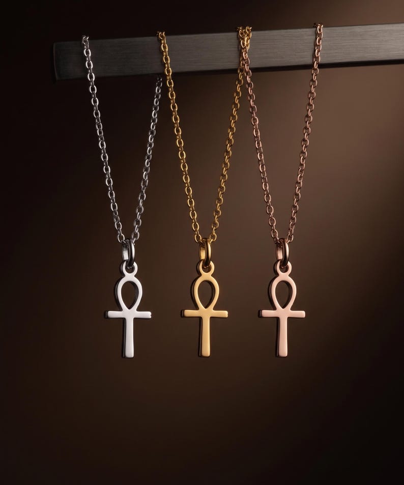 May include: Three Ankh pendant necklaces in silver, gold, and rose gold. Each necklace features a delicate chain and a stylized Ankh symbol, suspended against a dark brown background. The pendants are displayed hanging from a dark metal bar.