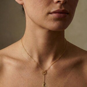 May include: A gold-colored necklace featuring a sun pendant at the top, with a chain extending down to a crescent moon charm. The necklace is worn around the neck, with the sun and moon pendants resting on the chest.