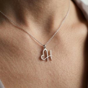 May include: A silver necklace with a heart-shaped pendant that includes the letter "H". The pendant hangs from a delicate chain. The necklace is worn around the neck.