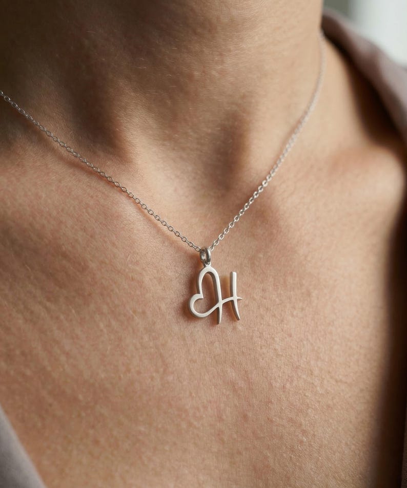 May include: A silver necklace with a heart-shaped pendant that includes the letter "H". The pendant hangs from a delicate chain. The necklace is worn around the neck.