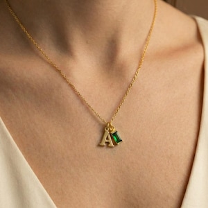 May include: Gold-colored necklace with a delicate chain. The pendant features the letter "A" and a rectangular emerald-green gemstone. The necklace is worn against a cream-colored top.