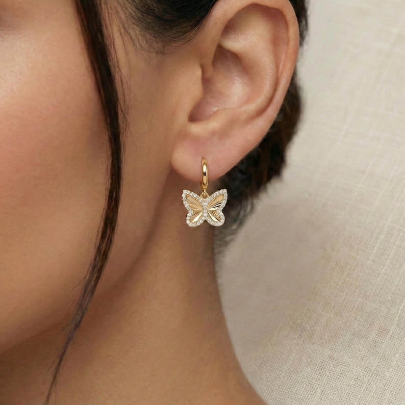 May include: A gold-colored butterfly earring with a small hoop. The butterfly has a textured design with a diamond-like border. The earring is worn on the ear of a person.