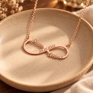 May include: A rose gold-toned infinity symbol necklace featuring the engraved names Susan and Mark along with the date 05.10.2022. The pendant is attached to a delicate matching chain, showcasing a polished metallic finish and cursive lettering.
