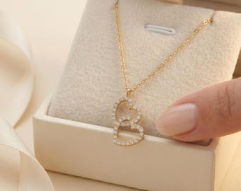 Puede incluir: Un collar dorado con dos colgantes entrelazados en forma de corazón, cada uno delineado con pequeñas piedras brillantes. El collar se exhibe en una caja de joyería abierta de color crema, con una cinta en el fondo.