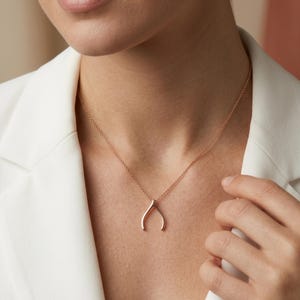 Op de afbeelding: Een roségouden wishbone-hangerketting. De hanger heeft een eenvoudig, gebogen ontwerp dat aan een delicate ketting hangt. De ketting wordt gedragen over een witte blazer.