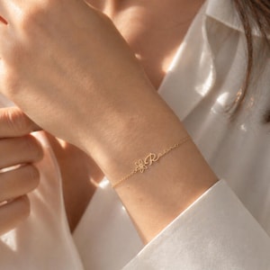 May include: A delicate gold bracelet with a floral design and the cursive letter "R". The bracelet is worn on a wrist, against a white button-down shirt. The bracelet's chain is thin and the overall aesthetic is minimalist and elegant.