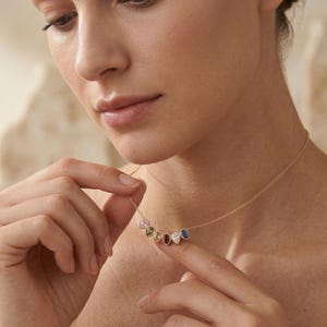 May include: A gold necklace featuring seven teardrop-shaped gemstones in various colors. The gemstones are arranged in a curved line, suspended from a delicate gold chain. The gemstones include pink, green, white, red, blue, and other colors.