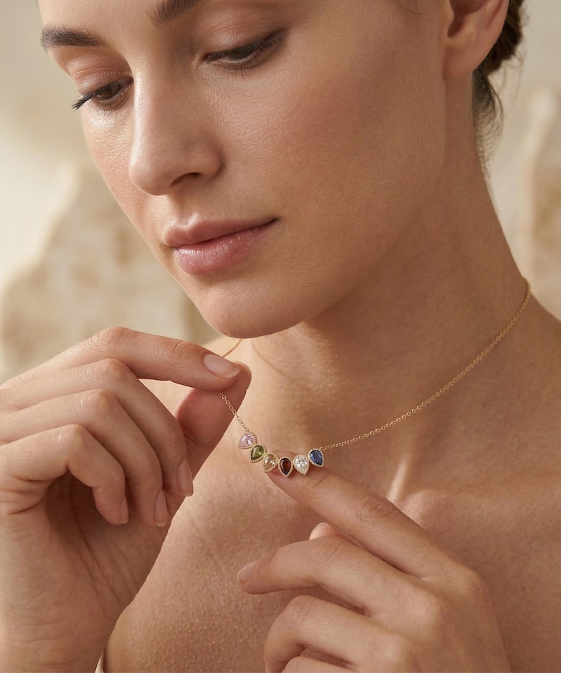 May include: A gold necklace featuring seven teardrop-shaped gemstones in various colors. The gemstones are arranged in a curved line, suspended from a delicate gold chain. The gemstones include pink, green, white, red, blue, and other colors.