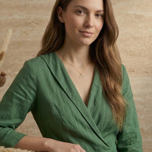 May include: A woman wearing a gold necklace with a pendant in the shape of the letter "H". The woman is wearing a green dress. The background is a neutral beige color.