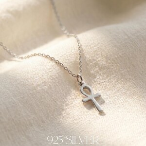 May include: A silver ankh pendant necklace. The ankh, a symbol of life, hangs from a delicate chain. The necklace is made of 925 silver and is displayed on a textured, off-white surface. The text "925 SILVER" is at the bottom.