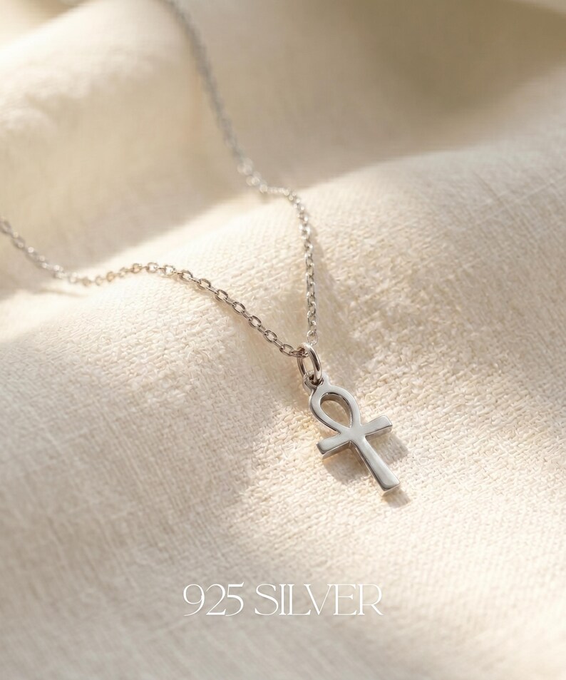 May include: A silver ankh pendant necklace. The ankh, a symbol of life, hangs from a delicate chain. The necklace is made of 925 silver and is displayed on a textured, off-white surface. The text "925 SILVER" is at the bottom.